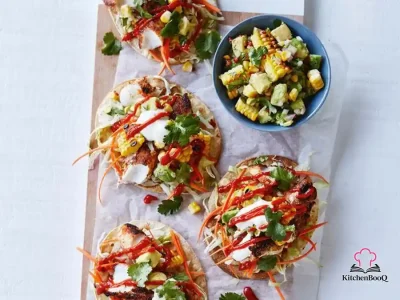 Crispy corn tortilla tostadas topped with flaky Mexican-seasoned flathead fish, charred corn and avocado salsa, shredded iceberg lettuce, and carrot matchsticks served on a white plate with lime wedges